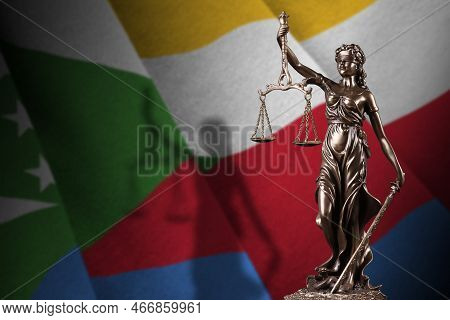 Comoros Flag With Statue Of Lady Justice And Judicial Scales In Dark Room. Concept Of Judgement And 