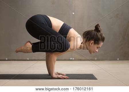 Young attractive smiling yogi woman practicing yoga, standing in Bakasana exercise, Crane pose, working out, wearing black sportswear, cool urban style, full length, grey studio background, side view