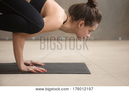 Portrait of young attractive smiling yogi woman practicing yoga, standing in Bakasana exercise, Crane pose, working out wearing black sportswear, cool urban style grey studio background, close up