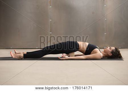 Young attractive sporty yogi woman practicing yoga, stretching in dvi pada pithasana exercise using wooden block, working out, wearing black sportswear, cool urban style, full length, grey studio