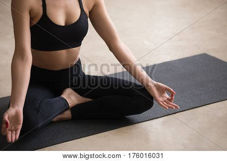 Young sporty yogi woman practicing yoga, sitting in Sukhasana exercise, Easy Seat pose with mudra, working out, wearing black sportswear, urban style grey studio floor background, midsection closeup