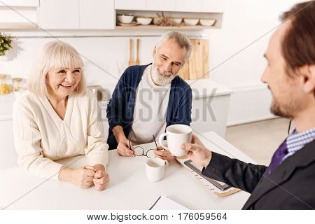 Enjoyable working hours. Amused glad cheerful notary having meeting and enjoying the hospitality of aged couple of clients while expressing holding the cup of tea