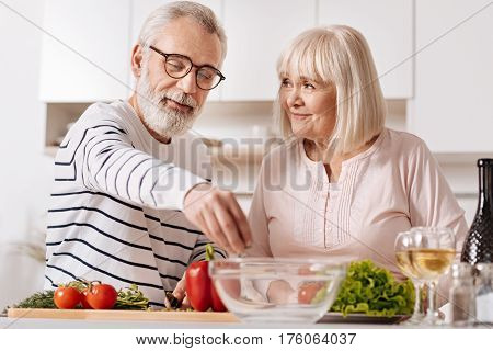 Enjoying healthy nutrition. Lively careful upbeat senior couple standing in the kitchen and cooking healthy dinner while expressing care and love