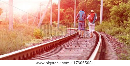 Two jolly Hikers Man and Girl in casual Travel Clothing with Backpacks walking along Railroad with back light Sun and Forest on Background