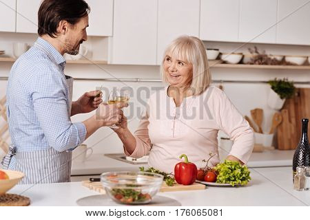 Time flies. Positive charismatic mature man standing in the kitchen and celebrating holiday while enjoying glass of wine with his aging mother