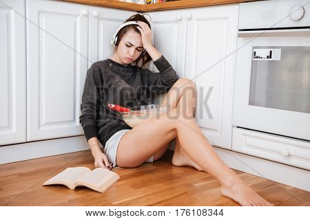 Portrait of an upset frustrated girl in headphones sitting on the kitchen floor and reading recipe book
