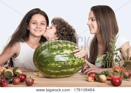 Children sit at the table in front of a large watermelon whole. Boy kisses girl on the cheek. White background. Close-up.