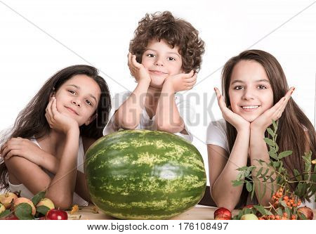 Two girls and a boy with his head on his hands sitting at the table in front of a large watermelon whole. Looking at the camera. White background. Close-up.