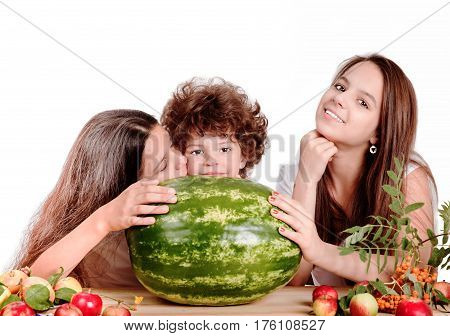 Brother and two sisters are sitting at the table in front of ripe watermelon. A girl kisses a boy on the cheek. White background. Close-up.