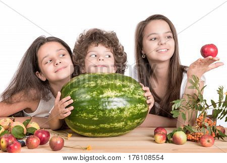 Children sit at a wooden table and play with fruit. Before them lies a large watermelon. White background. Close-up.