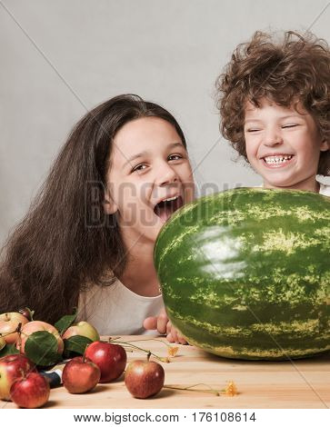 Brother and sister playing with a huge whole watermelon. The girl wants to take a bite of watermelon. Boy laughs. Gray background. Close-up.
