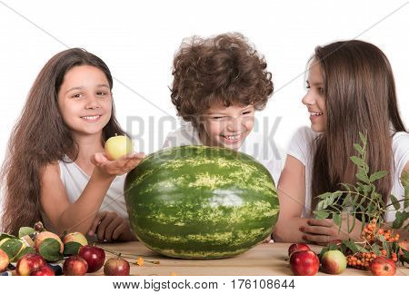 Brother and two sisters laughing while sitting at the table. On the table is a huge whole watermelon and fruit. White background. Close-up.