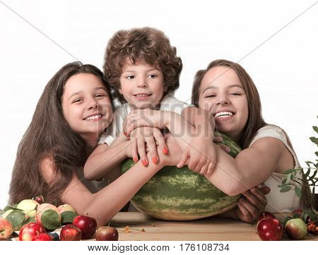Three happy children hugging a huge watermelon sitting at the table. Smiling looking into the camera. White background. Close-up.