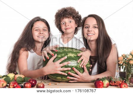 Three happy children holding hands on a huge watermelon sitting at the table. Smiling looking into the camera. White background. Close-up.