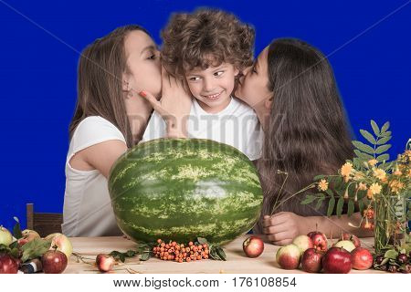 The sisters whisper in the ear of the little brother Curly. Before them lies a huge whole watermelon and fruit. Close-up.