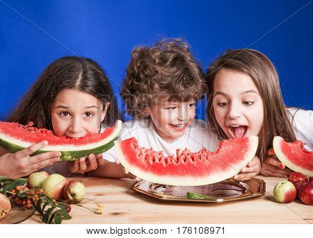 Little curly boy and two girls sitting at a wooden table and eat large ripe watermelon. Blue background. Close-up.