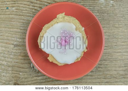 Single freshly baked homemade cookie decorated with a pink candy heart on white icing served on an orange plate viewed from overhead