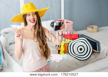 Young woman making selfie portrait with hat in the bedroom with suitcase and clothes on the background. Prepairing for the summer vacations