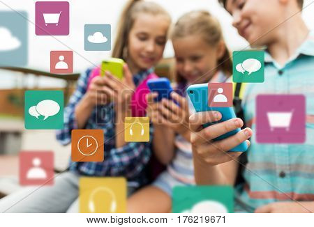primary education, friendship, childhood, technology and people concept - group of happy elementary school students with smartphones and backpacks outdoors over multimedia icons