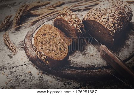 Bakery and grocery concept background. Fresh, healthy whole grain sliced bread loaf, sprinkled flour, wheat ears and knife cutting it on rustic table and wooden round desk, food closeup. Dark filtered