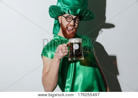 Drunk screaming man in st.patriks costume and eyeglasses which holding cup of beer over gray bakground