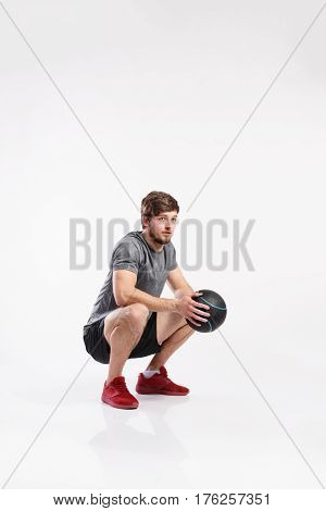 Handsome hipster fitness man in gray t-shirt holding medicene ball, crouching. Studio shot on gray background.