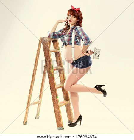 Fashionable redhead pregnant woman painter in pin up style with brush and ladder