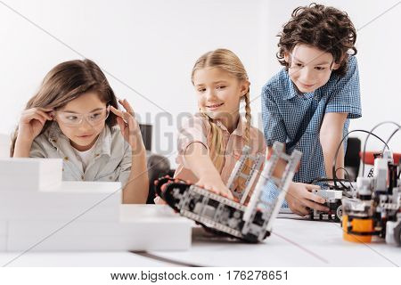 Robot test. Attentive joyful motivated kids sitting at school and testing cyber robots while working on the tech project