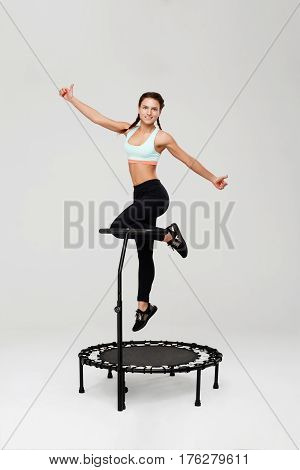 Young woman on mini rebounder jumping up showing thumbs up with smile isolated on grey