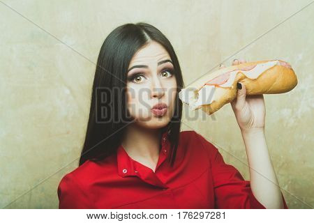 sexy cute woman or pretty girl with brunette long hair and makeup on surprised face in fashionable red shirt holds eats big burger or sandwich with sausage and cheese on textured wall background