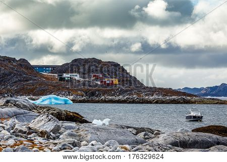 Stone Arctic Coast, Motorboat And Blue Iceberg Floating In The Bay With Small Colorful Houses On The