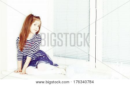 Little girl in blue striped dress and a white bow on her head.She sits at the window turned away from the camera.Creative toning of a photograph.