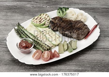 Grilled beef grilled with asparagus, zucchini, lobi, broccoli, tomato, chili and red sauce on a white plate, over wood background