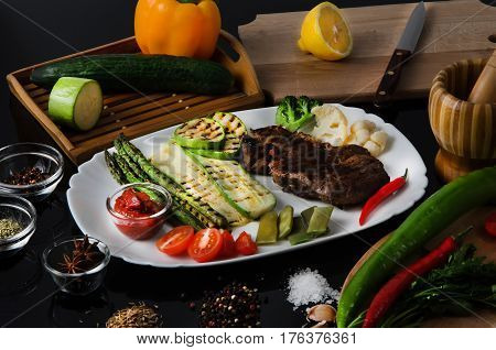 Grilled beef grilled steak with asparagus, zucchini, lobi, broccoli, tomato, chili and red sauce on a white plate, on a black glossy background