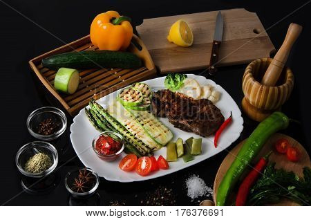 Grilled beef grilled steak with asparagus, zucchini, lobi, broccoli, tomato, chili and red sauce on a white plate, on a black glossy background