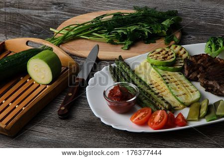 Grilled beef grilled with asparagus, zucchini, lobi, broccoli, tomato, chili and red sauce on a white plate, and other vegetables in wood background