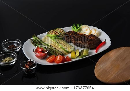 Grilled beef grilled steak with asparagus, zucchini, lobi, broccoli, tomato, chili and red sauce on a white plate, on a black glossy background