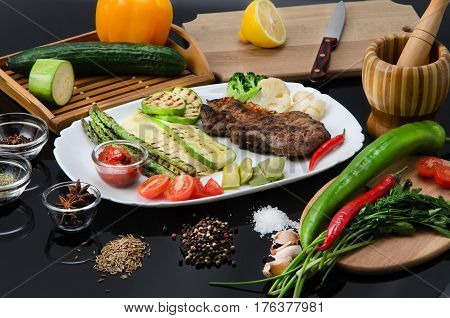 Grilled beef grilled steak with asparagus, zucchini, lobi, broccoli, tomato, chili and red sauce on a white plate, on a black glossy background
