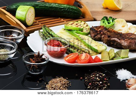 Grilled beef grilled steak with asparagus, zucchini, lobi, broccoli, tomato, chili and red sauce on a white plate, on a black glossy background
