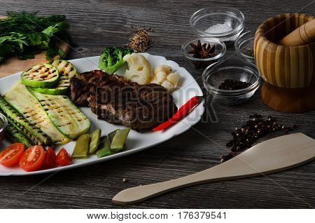 Grilled beef grilled with asparagus, zucchini, lobi, broccoli, tomato, chili and red sauce on a white plate, and other vegetables in wood background