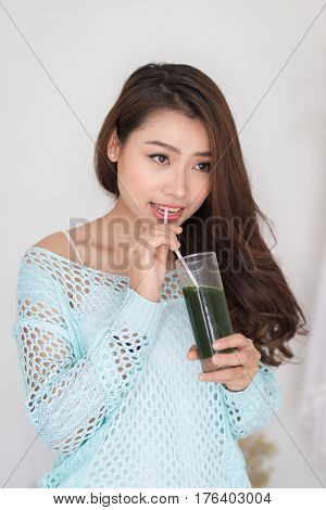 Smiling young asian woman drinking green fresh vegetable juice or smoothie from glass at home