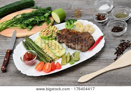 Grilled beef grilled with asparagus, zucchini, lobi, broccoli, tomato, chili and red sauce on a white plate, and other vegetables in wood background
