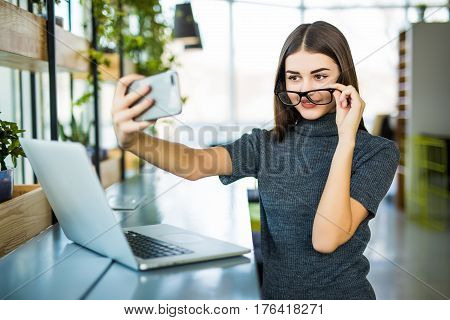 Young female taking a selfie in the office while sitting with laptop in modern office room