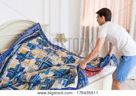 handsome young man making bed a color patchwork quilt in the white interior. scrappy blanket on the bed closeup.