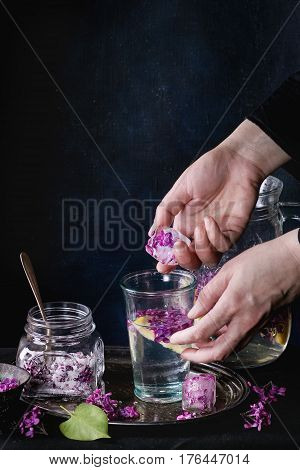 Femail hands put ice cubes with flowers into glass of lilac lemonade with lemon. Glass jar of sugared lilac flowers and pitcher on black tablecloth over black. Dark rustic atmosphere. See series.