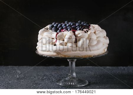 Homemade meringue cake Pavlova with whipped cream, fresh blueberries and blueberry sauce on vintage cake stand on black concrete texture background