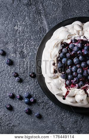 Homemade meringue cake Pavlova with whipped cream, fresh blueberries and blueberry sauce on vintage cake stand on black concrete texture background. Top view, space