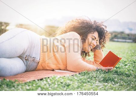 Cheerful young woman with curly hair lying on the ground in a park on sunny day reading book with pleasure - Smiling girl holding her diary while relaxing on the grass - Education and culture concept