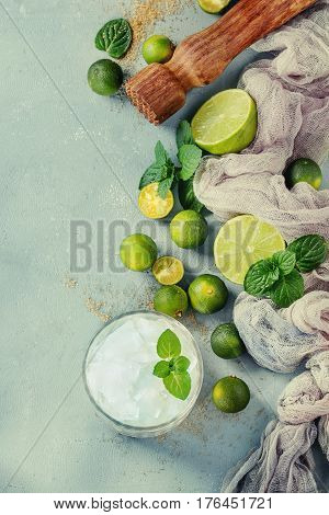 Ingredients for mojito cocktail, whole, sliced lime and mini limes, mint leaves, brown crystal sugar over gray stone texture background, gauze textile, bar muddler, glass of ice. Top view, space