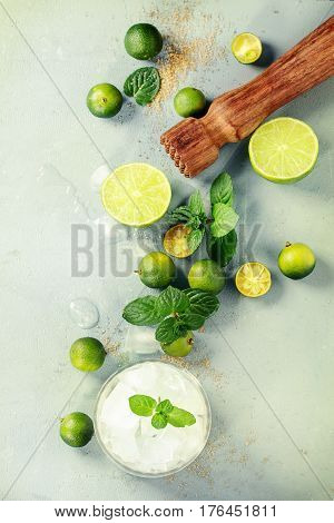 Ingredients for mojito cocktail, whole, sliced lime and mini limes, mint leaves, brown crystal sugar over gray stone texture background with wooden bar muddler, glass full of ice. Top view, space